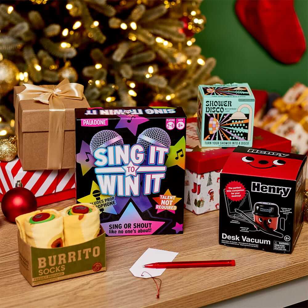 Selection of novelty Christmas gifts, including a singing game, desk vacuum, shower disco gadget and burrito socks, displayed on a table in front of a decorated Christmas tree. Selection of novelty Christmas gifts, including a singing game, desk vacuum, shower disco gadget and burrito socks, displayed on a table in front of a decorated Christmas tree.